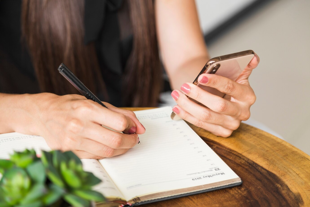 Mulher escreve em uma agenda e celular na mão